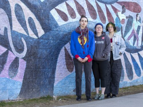 Sam Wallace, Brandi Tucker and Em Schluter pose against a wall mural outside DelSesto Middle School in Providence. Wallace, left, a librarian, and Schluter, right, an ESL teacher, both work at the school but were recently issued nonrenewal notices, which would prevent them from returning to teach in Providence next year. Tucker, also one of the 17 educators issued a nonrenewal this year in the Providence public school system, works at nearby Webster Avenue Elementary School. The educators are appealing their nonrenewals.